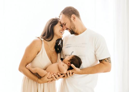Two happy new parents closely embracing one another in a light setting while holding a naked newborn baby